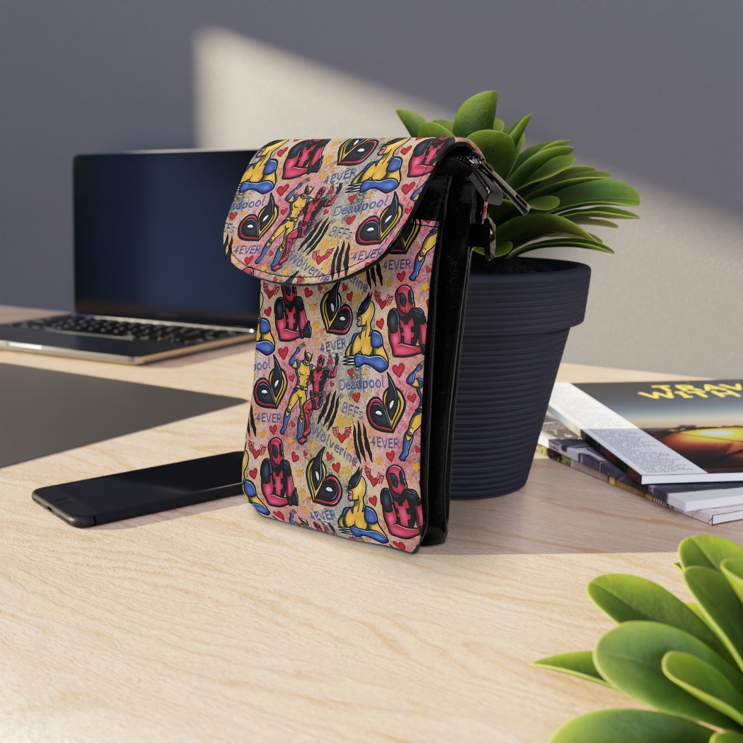 Deadpool x Wolverine Cell Phone Wallet Bag with colorful comic design sitting on desk near plant and electronic devices
