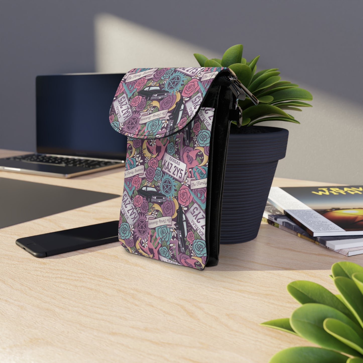 Supernatural Winchester Brothers Floral Cell Phone Wallet Bag displayed on a light wood desk near a plant and laptop.