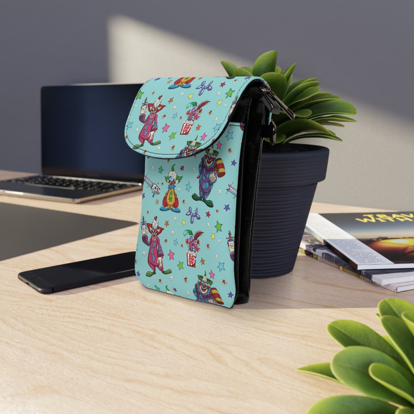Killer Klowns Cell Phone Wallet Bag with colorful clown design displayed on a wooden desk near plants and books