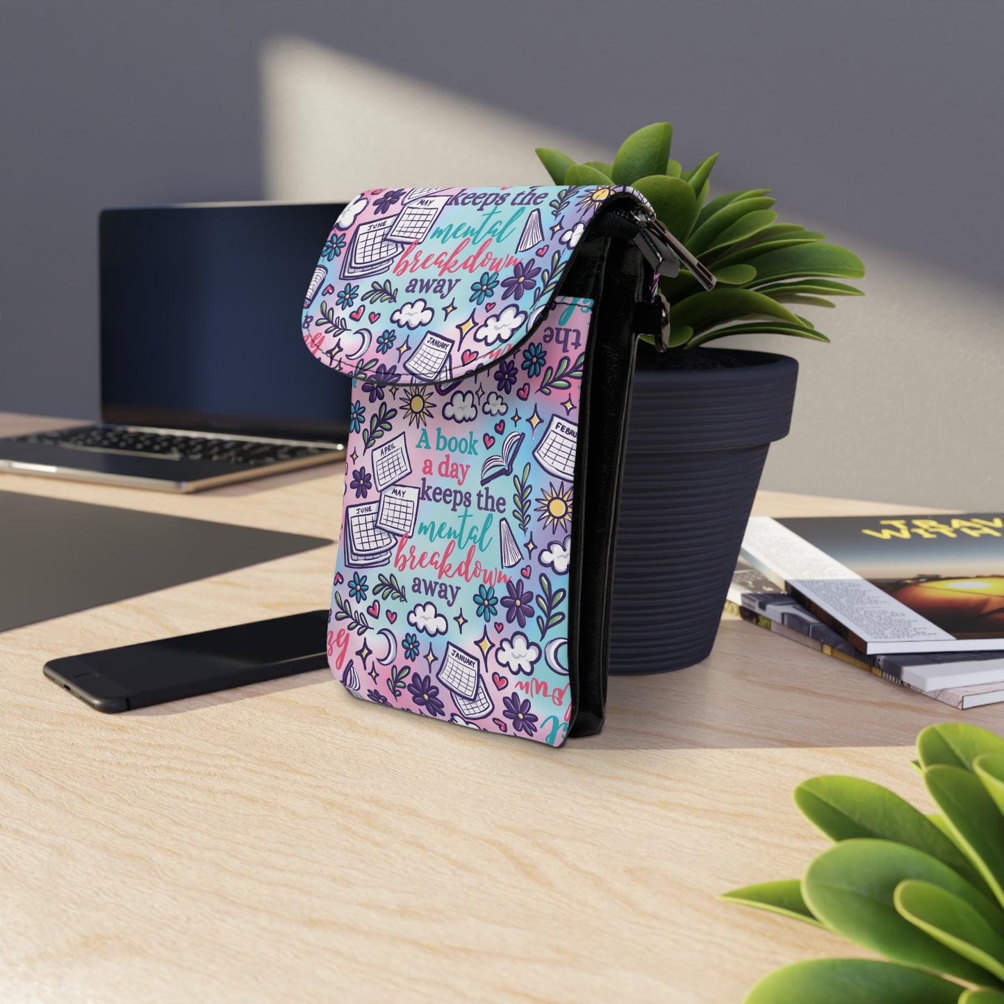 A colorful A Book A Day Keeps The Mental Breakdown Away Cell Phone Wallet Bag standing on a desk near plants and a laptop