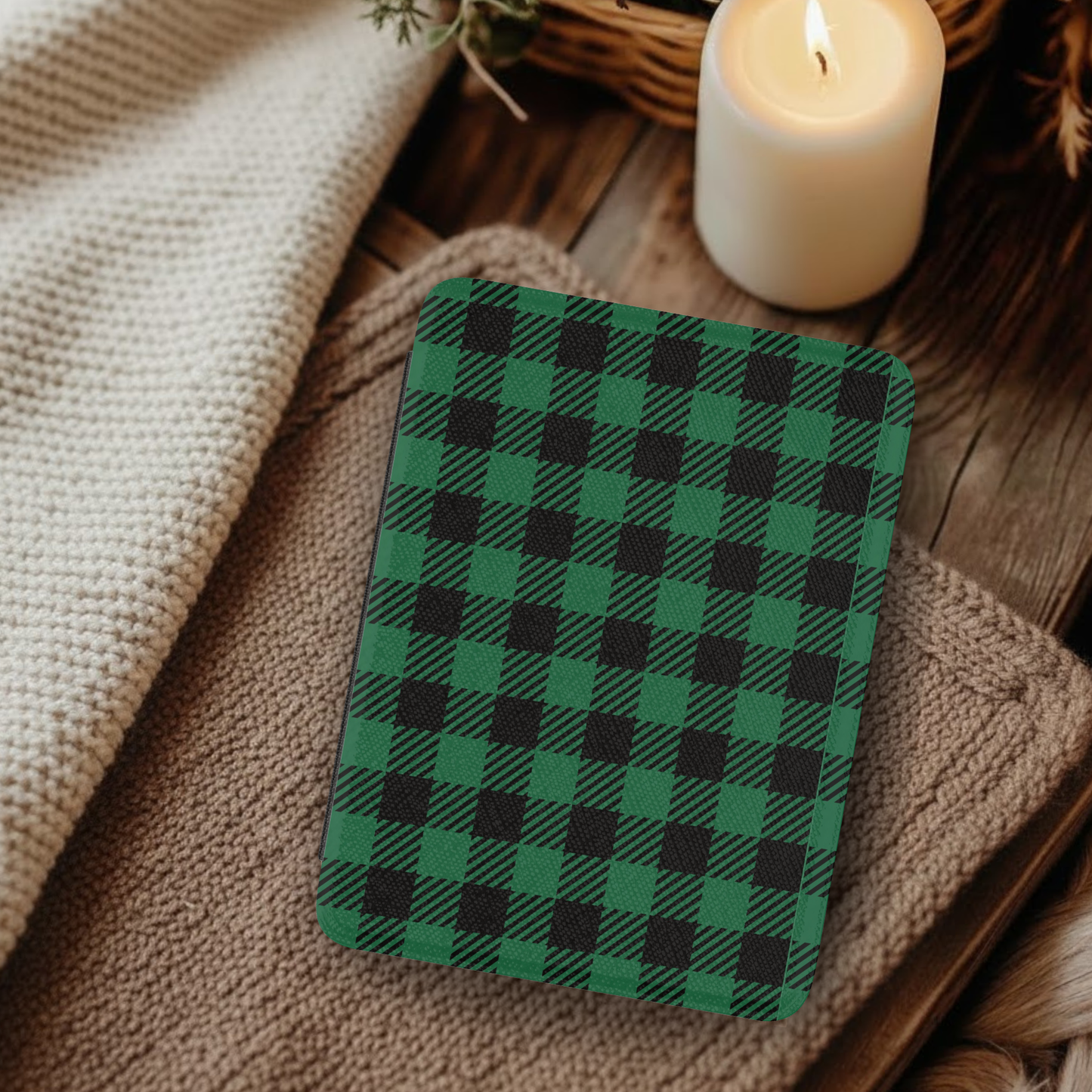 Green Buffalo Plaid Leather Kindle Case with Hand Strap resting on cozy knit blankets beside a lit candle on wooden surface