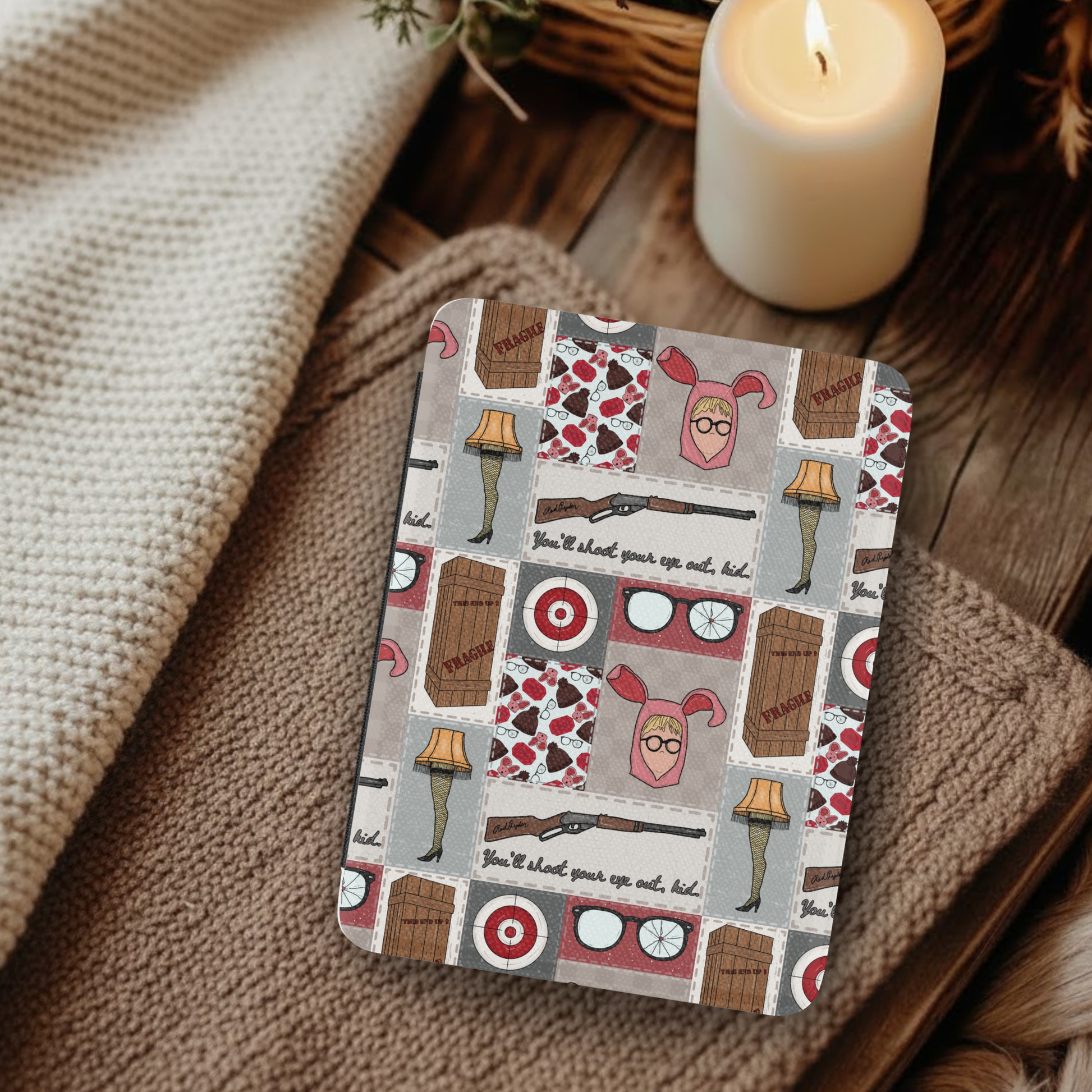 A Christmas Story Patchwork Leather Kindle Case with Hand Strap displayed beside cozy blankets and a lit candle on wood.