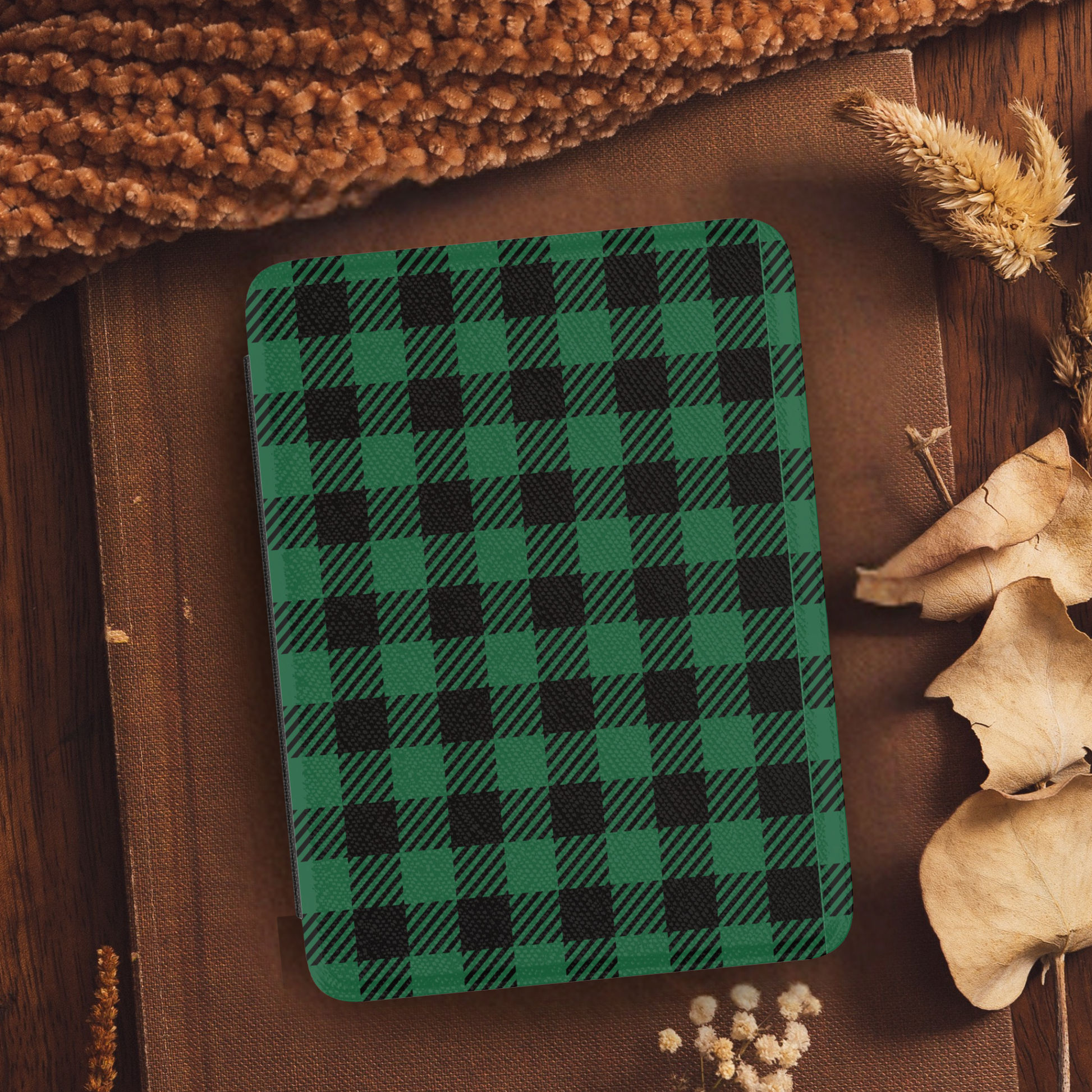 Green Buffalo Plaid Leather Kindle Case with Hand Strap resting on rustic brown fabric with dried leaves and plants nearby
